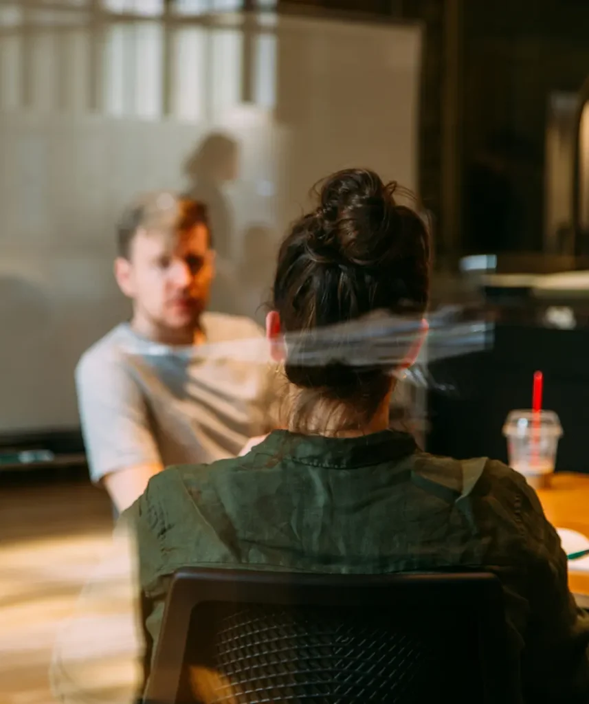 Genom en glasruta ser vi två personer diskutera i ett mötesrum.